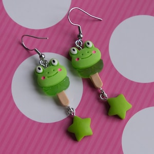 May include: Green frog popsicle earrings with a green star charm. The frog has pink cheeks and a white stick.