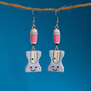 May include: A pair of blue and pink pencil sharpener earrings. The earrings are made of polymer clay and feature a smiling face on the front. The earrings are hanging from silver hooks.