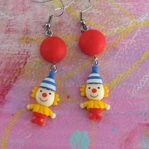 May include: A pair of dangle earrings featuring a red ball and a cartoon clown with a yellow shirt, blue and white striped hat, and red pants.