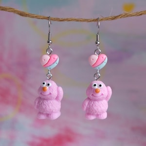 Puede incluir: Pendientes de monstruo peludo rosa con un dije de corazón rosa, azul y blanco.