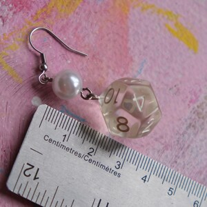 May include: A pair of dangle earrings featuring a clear plastic dice charm with the numbers 8 and 10 visible, and a white pearl bead. The earrings are measured against a ruler in centimeters.