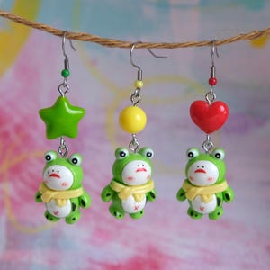 May include: Three pairs of novelty earrings hanging from a brown string. Each earring features a green frog with a yellow scarf. One pair has a green star, another a yellow bead, and the last a red heart.