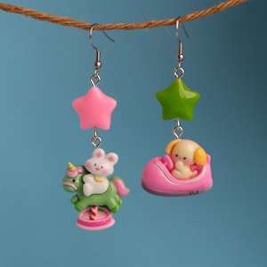 May include: A pair of dangle earrings with a pink star charm and a green unicorn charm with a white bunny riding it on one earring and a green star charm and a pink car with a yellow dog inside on the other earring.