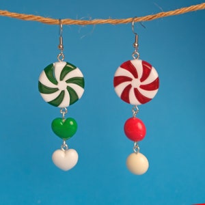 May include: A pair of dangle earrings featuring a peppermint candy design. One earring has a red and white peppermint candy, the other has a green and white peppermint candy. Both earrings have a white heart charm and a red or green heart charm.