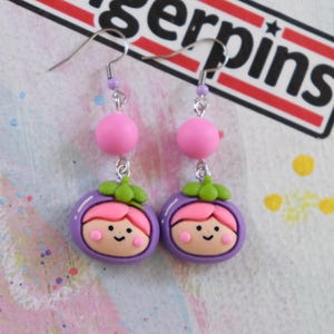 May include: Dangle earrings with a purple mangosteen design, featuring a smiling face and pink hair. The earrings have pink beads and silver hooks. Handmade earrings.