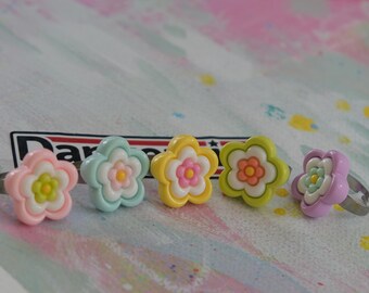 Op de afbeelding: Vijf bloemvormige ringen in pastelkleuren: roze, lichtblauw, geel, groen en paars. Elke ring heeft een wit midden met een contrasterende kleur in het midden. De ringen hebben zilverkleurige verstelbare bandjes.