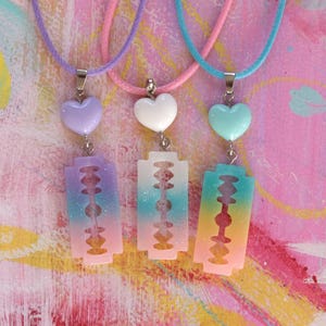May include: Three necklaces with pastel-coloured heart charms and rainbow-coloured razor blade pendants. The necklaces are on a pink and yellow background.