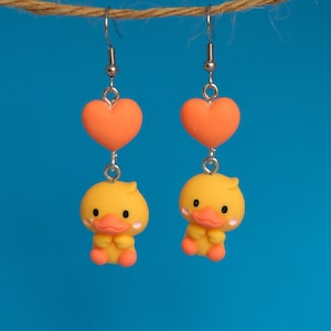 May include: A pair of dangle earrings featuring a yellow duck charm and a coral heart charm. The earrings are made of polymer clay and have silver hooks.