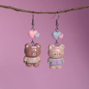 May include: A pair of dangle earrings featuring brown teddy bears wearing pastel colored jackets and holding pink and blue hearts.
