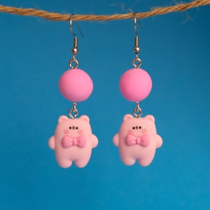 May include: Pink dangle earrings featuring a small pink bear with a bow tie and a pink ball charm.