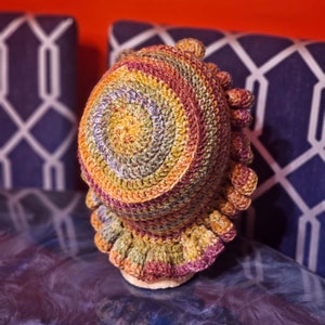 May include: A handmade crochet hat in a spiral pattern of orange, yellow, green, and purple. The hat has a ruffled brim and a textured, rounded crown. The hat is displayed on a light-colored surface.
