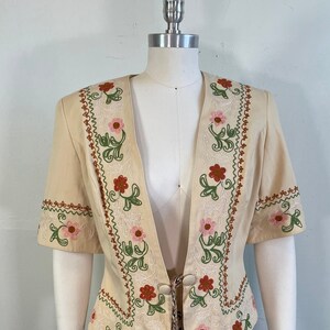 May include: A cream-colored embroidered jacket with a V-neck and short sleeves. The jacket features a floral design in shades of pink, red, green, and brown. The jacket has a tie at the waist.