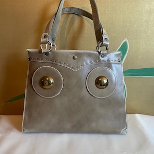 May include: A light brown leather tote bag with two large gold circular accents on the front. The bag has two top handles and a stitched design.
