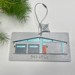 May include: A rectangular, glittery gray ornament featuring a house design with teal and red accents. The ornament has the text "OUR HOME" and "2023". A silver string is attached for hanging. A sprig of evergreen is in the background.