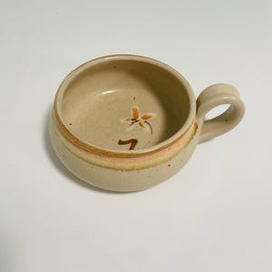 May include: A beige ceramic soup bowl with a handle. The bowl has a floral design and a brown and orange stripe around the rim. The bowl is round and has a handle for easy use.