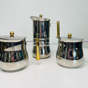 May include: Three stainless steel coffee pots with gold-colored handles and knobs. The set includes a traditional stovetop espresso maker and two smaller pots, all with a shiny, reflective surface. The coffee pots are arranged on a white surface.