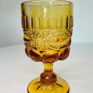 May include: Amber-colored vintage goblet with ornate detailing. The glass features a textured design around the bowl and a decorative stem. The goblet has a round base and is likely used for serving beverages.