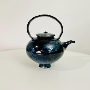 May include: A ceramic teapot with a dark blue and black glaze. The teapot has a rounded body, a small spout, and a curved handle. The lid sits atop the teapot, and the base is a small pedestal. The teapot is a decorative item.