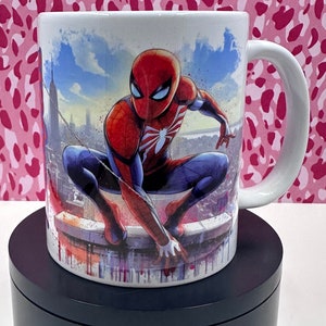 May include: White ceramic mug with a colorful illustration of Spider-Man in a superhero pose. The background is a cityscape with a blue sky.