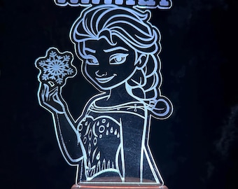 Könnte beinhalten: Eine leuchtende LED-Nachtlampe mit einem Cartoon-Bild einer Prinzessin, die eine Schneeflocke hält, mit dem Namen "NIHIRA" darüber. Die Lampe hat einen Holzsockel und strahlt ein weiches, weißes Licht aus, ideal für ein Kinderzimmer.