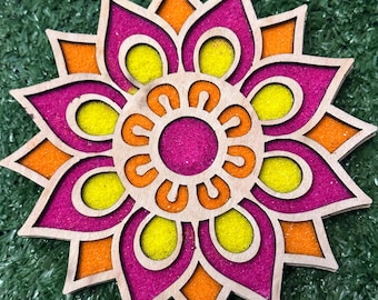 Puede incluir: Un mandala de madera con un diseño rosa, naranja y amarillo. El mandala está lleno de arena.
