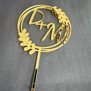 May include: Gold cake topper with a double circle design and the letters "D + M" in the centre. The circle is decorated with gold leaves.