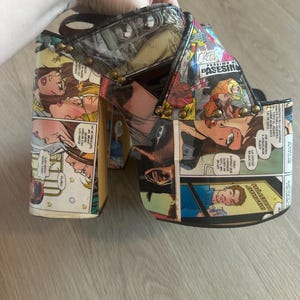 May include: Platform heels with a comic book design. The shoes feature a thick platform sole and a chunky heel. The upper part of the shoe is covered in panels from a comic book, with images of people and speech bubbles.