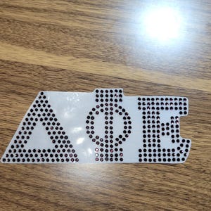 May include: A white sticker with the letters "AΦE" in black rhinestones. The letters are in a stylised font, with the "A" being a triangle and the "E" being a stylised letter.