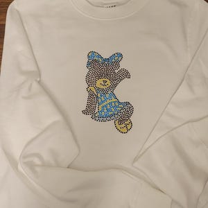 May include: White long-sleeved shirt with a rhinestone bear design. The bear design features blue, yellow, and black rhinestones. The shirt has a crew neck and long sleeves. The design is centered on the chest.