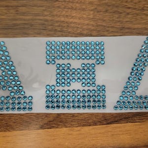 May include: A blue rhinestone design of the letters "AXA" on a white background.