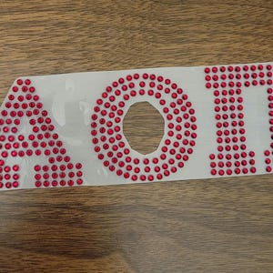 May include: A white decal featuring the letters AOI, each letter is decorated with small red rhinestones. The decal is placed on a wooden surface.