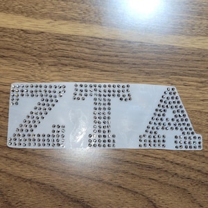May include: A white patch with the letters "ZTA" spelled out in silver rhinestones.