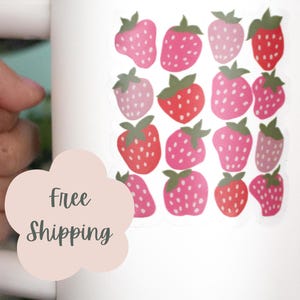 May include: White travel mug decorated with a pattern of pink strawberries with green leaves. The mug has a white handle and a white lid. The text "Free Shipping" is on a light pink flower-shaped sticker.