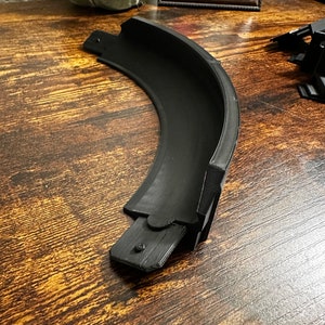 May include: A black plastic track piece for a toy race car set. The piece is curved and has a raised edge.