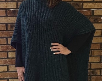 May include: A dark green knitted poncho with a turtleneck collar. The poncho is worn over a black mock neck shirt and black pants.