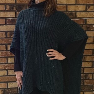 Op de afbeelding: Een donkergroene gebreide poncho met een rolkraag. De poncho wordt gedragen over een zwart shirt met een hoge hals en een zwarte broek.