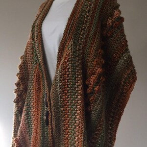May include: A handmade, open-front crochet cardigan in shades of brown, green, and orange. The textured knitwear features a vertical striped pattern and decorative bobbles along the sleeves. The garment is displayed on a white form.