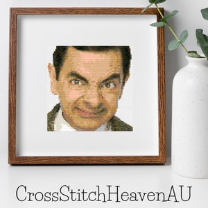 Op de afbeelding: Kruissteekpatroon van Mr. Bean, een populair Brits komisch personage, met een close-up van zijn gezicht. Het patroon is in een bruine houten lijst.