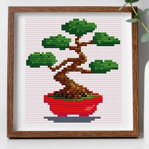 May include: Cross stitch pattern of a bonsai tree in a red pot. The tree has a brown trunk and green leaves. The pattern is framed in a brown wooden frame.