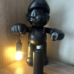 Puede incluir: Escultura negra de un personaje de dibujos animados sosteniendo una bombilla y de pie junto a una lata de pintura en aerosol. La escultura está sobre una superficie de madera.