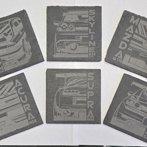 May include: Six dark gray slate coasters with etched car designs. Each coaster features a different car model with the car's name and model number. The coasters are square and have a rough texture.