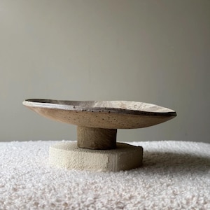 Peut inclure: Un bol en céramique beige moucheté peu profond avec un bord large et irrégulier, soutenu par une base cylindrique marron qui repose sur un bloc rectangulaire épais, texturé et blanc cassé, formant un objet décoratif à plusieurs niveaux.