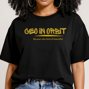 Könnte beinhalten: Schwarzes T-Shirt mit dem goldenen Schriftzug "CEO IN ORBIT" und dem Slogan "Be your own kind of beautiful". Das Shirt hat einen Rundhalsausschnitt.