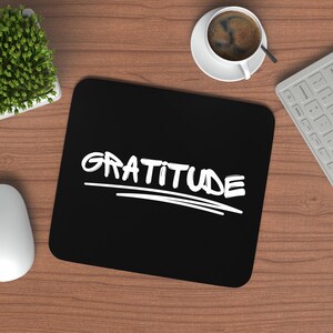 May include: A black mousepad with the word "GRATITUDE" in white script. The mousepad is on a wooden desk, with a cup of coffee, a keyboard, a mouse, and a small plant in the background.