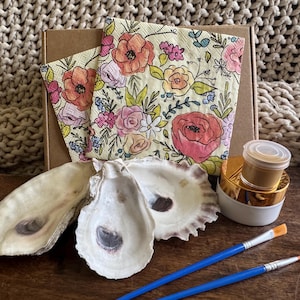 May include: An art kit with oyster shells, paint, and brushes. The kit includes two oyster shells, two paintbrushes, a small jar of gold paint, and floral napkins with pink and orange flowers.