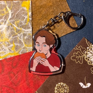 May include: A keychain featuring a cartoon illustration of a person with brown hair eating a hamburger. The person is wearing a red shirt. The keychain is made of clear acrylic.