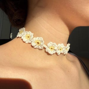 May include: A handmade crochet choker necklace featuring white daisy-like flowers with yellow centers. The necklace is worn around the neck and is made of a soft, textured yarn. The floral design adds a delicate and feminine touch.