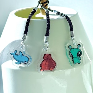 May include: Three clear acrylic charms with black braided cords. One charm is a blue cartoon shark, another is a red bear, and the third is a green alien with large eyes. Each charm has a silver-colored clasp.