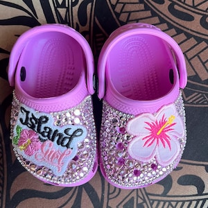 Puede incluir: Un par de zuecos rosas para niños con pedrería y diseños bordados. Un zueco presenta una flor de hibisco rosa y el otro un parche blanco con el texto "Island Girl".