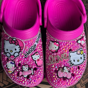 Pode incluir: Sandálias rosa com tema Hello Kitty com strass e pingentes Hello Kitty. As sandálias têm uma base rosa com personagens Hello Kitty e as palavras "Hello Kitty" na frente.